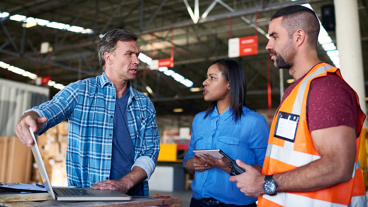 Three warehouse workers discussing operations, one in plaid shirt gesturing at laptop, another in blue shirt with tablet.