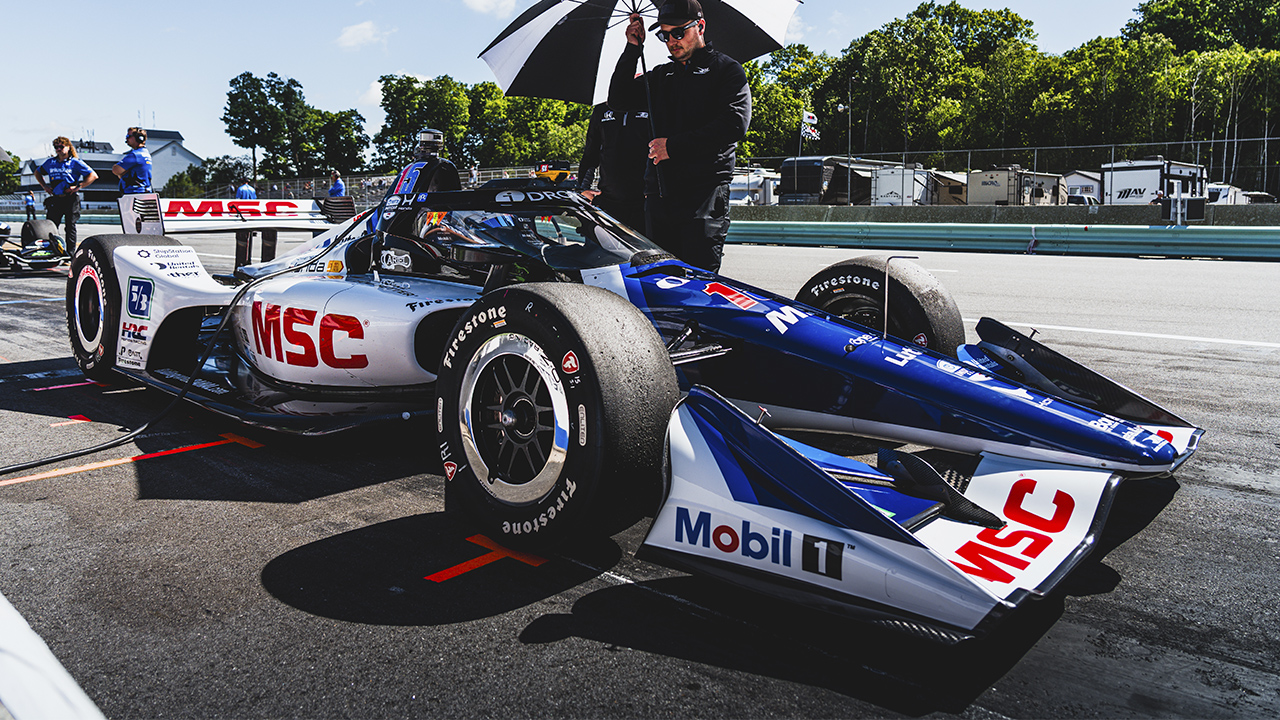 MSC-branded IndyCar race car in blue and white livery with Firestone tires on pit lane with crew member holding umbrella