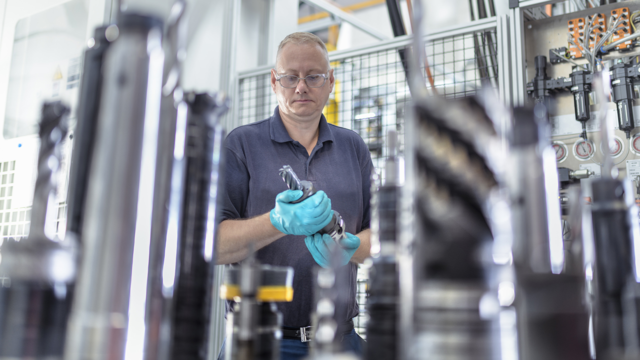Factory worker in glasses and blue gloves inspecting metal component among industrial machinery and equipment.