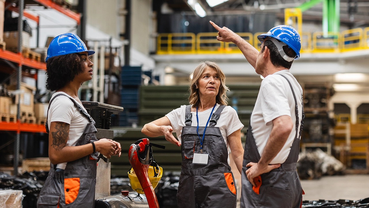Three warehouse workers in blue hard hats and gray overalls discussing operations in an industrial facility.