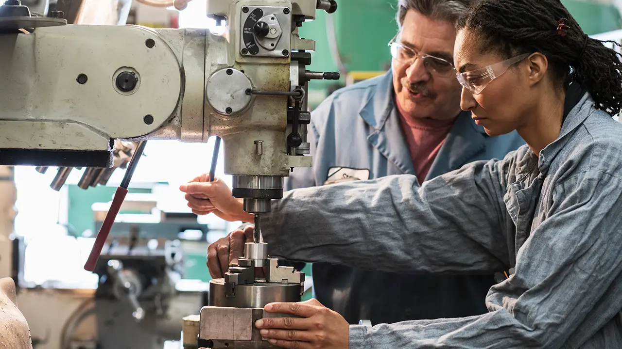 Two workers in safety glasses operating a drill press machine in an industrial workshop setting.