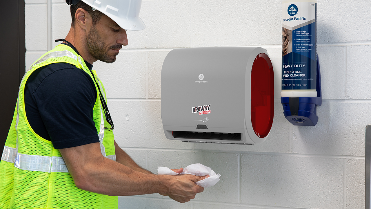Worker in safety vest and hard hat using wall-mounted paper towel dispenser and hand cleaner in industrial setting.