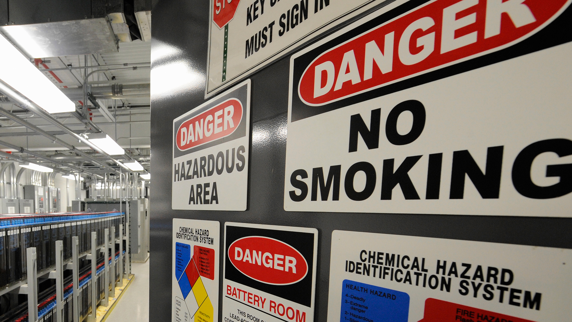 Multiple danger warning signs on a gray pillar in an industrial facility, including hazardous area, no smoking, and battery room warnings.