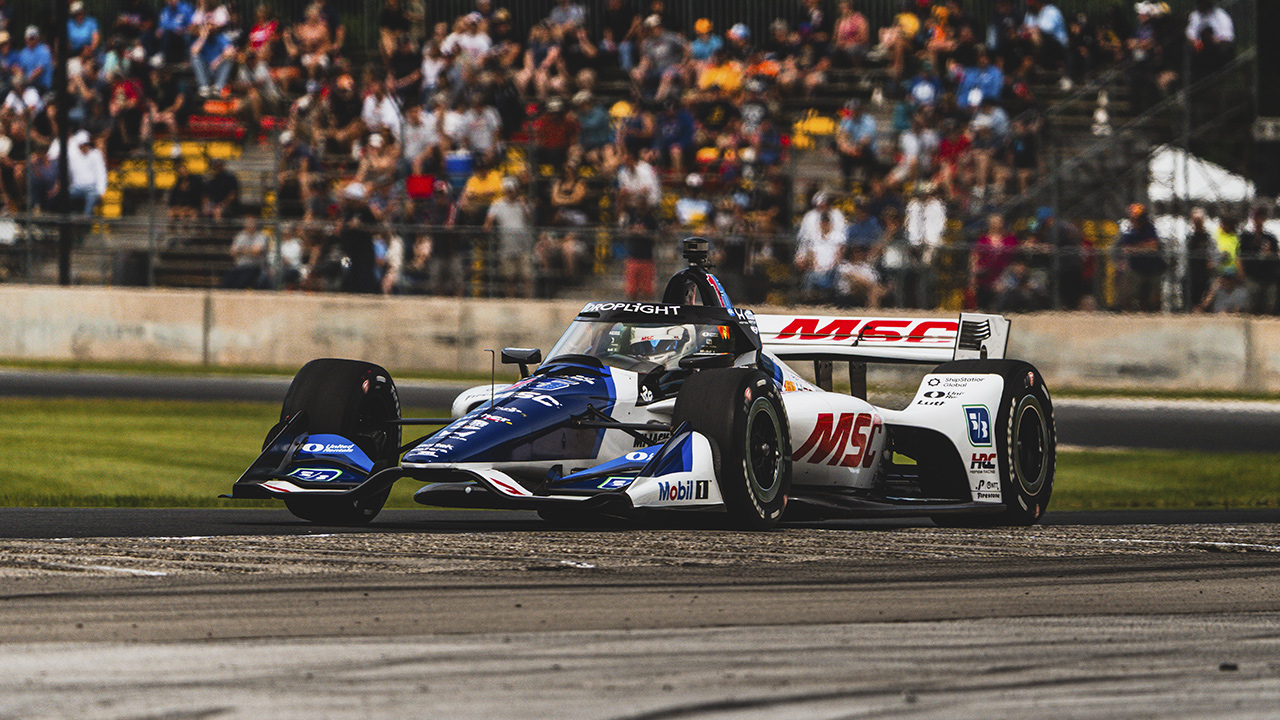 MSC-branded white, blue, and black IndyCar racing on track with crowded grandstands in background