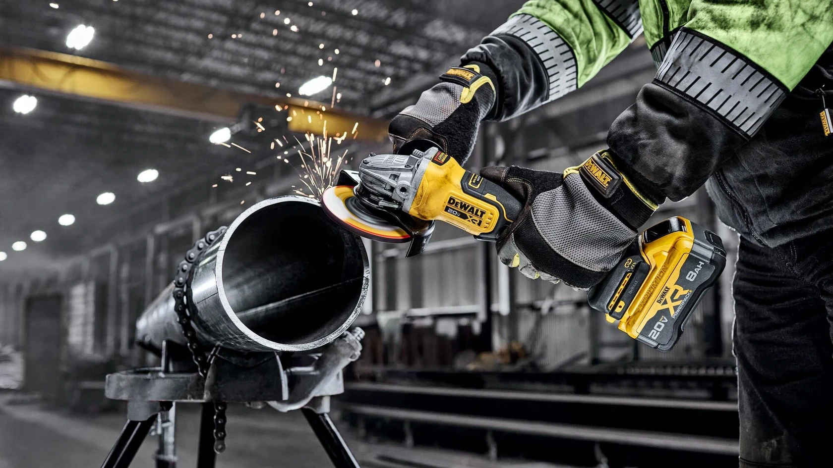 Worker in green jacket using yellow DeWalt angle grinder on metal pipe with sparks flying in industrial warehouse