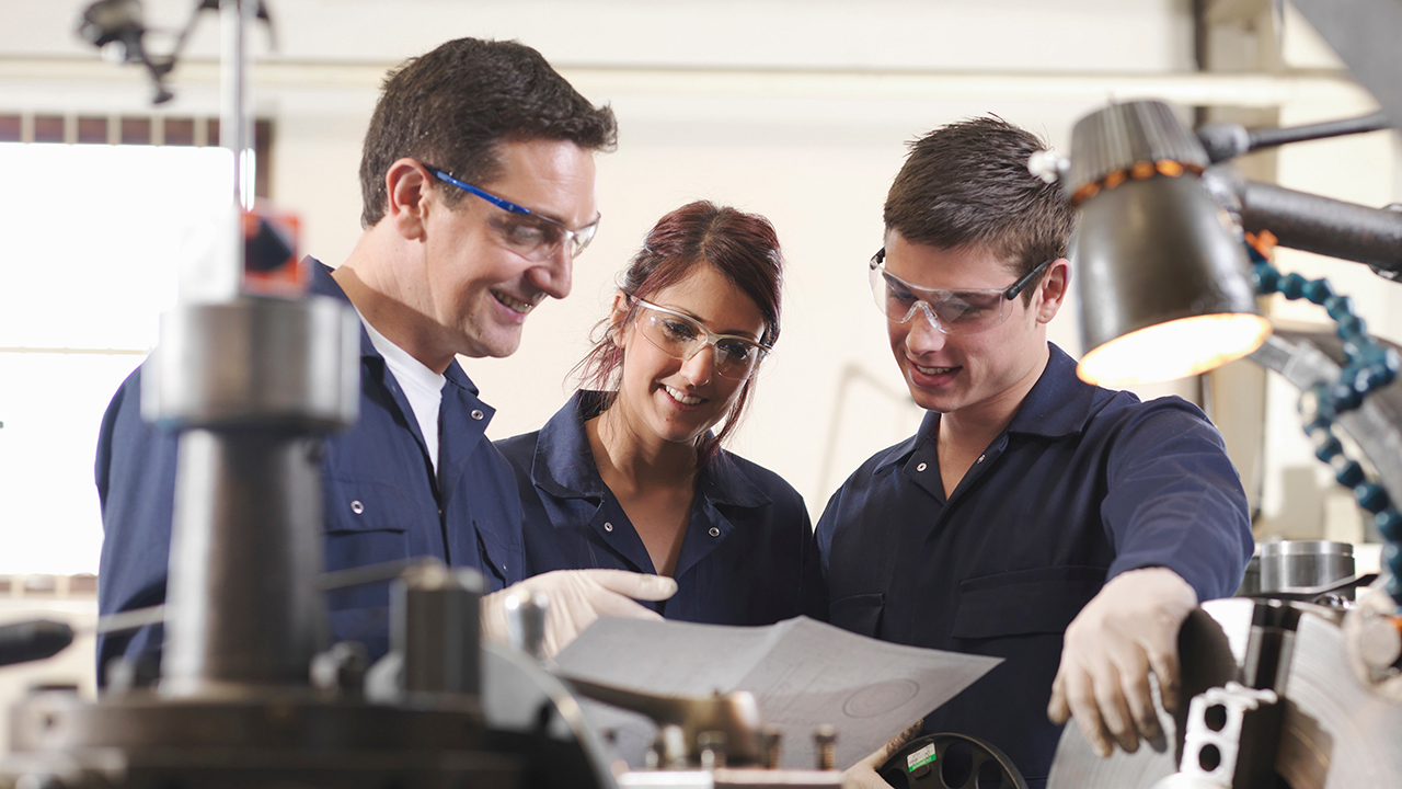 Three workers in navy uniforms and safety goggles examining a metal part together in a workshop with machinery.