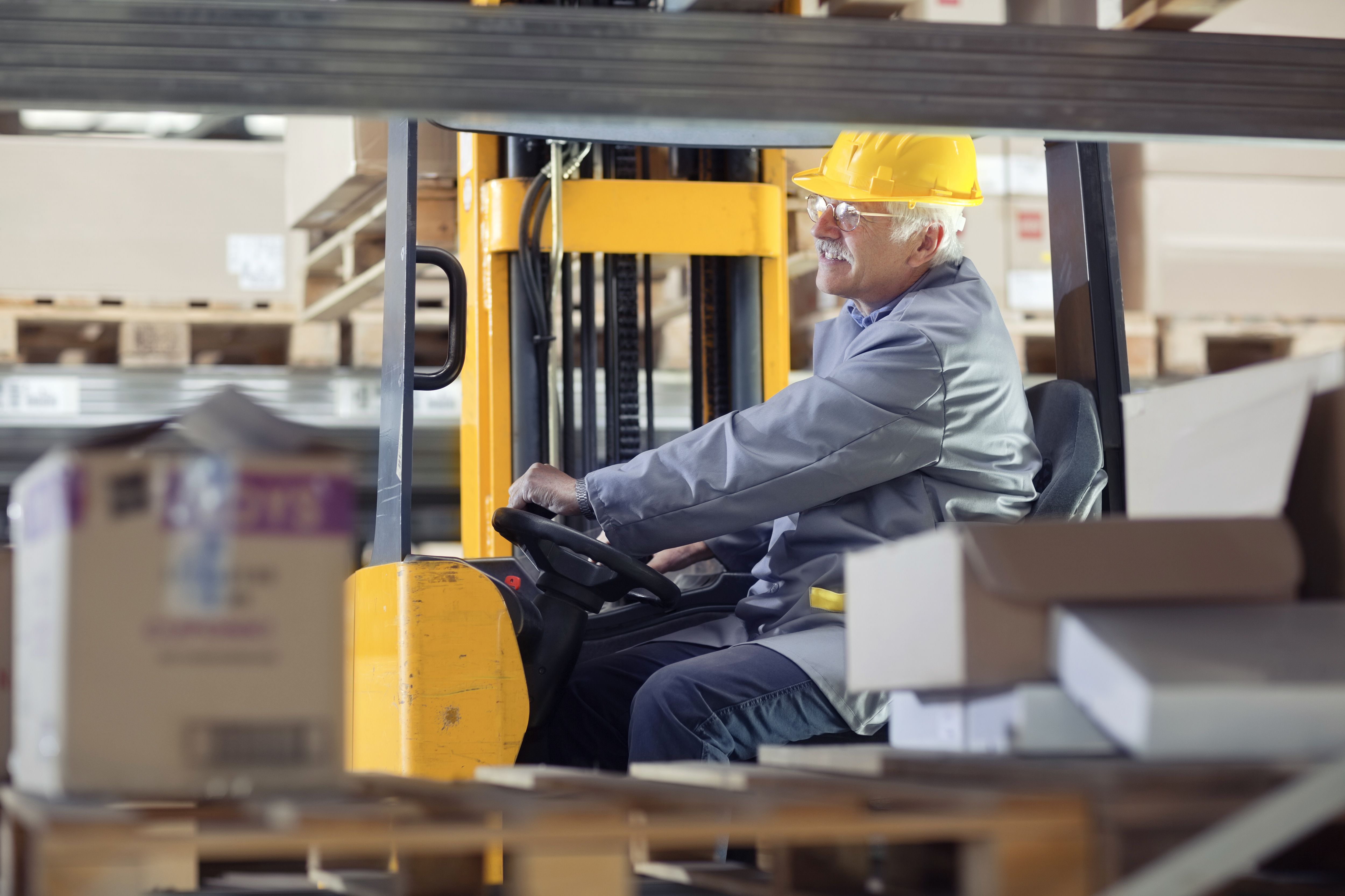 Worker in yellow hard hat operating yellow forklift in warehouse with wooden pallets and storage shelves