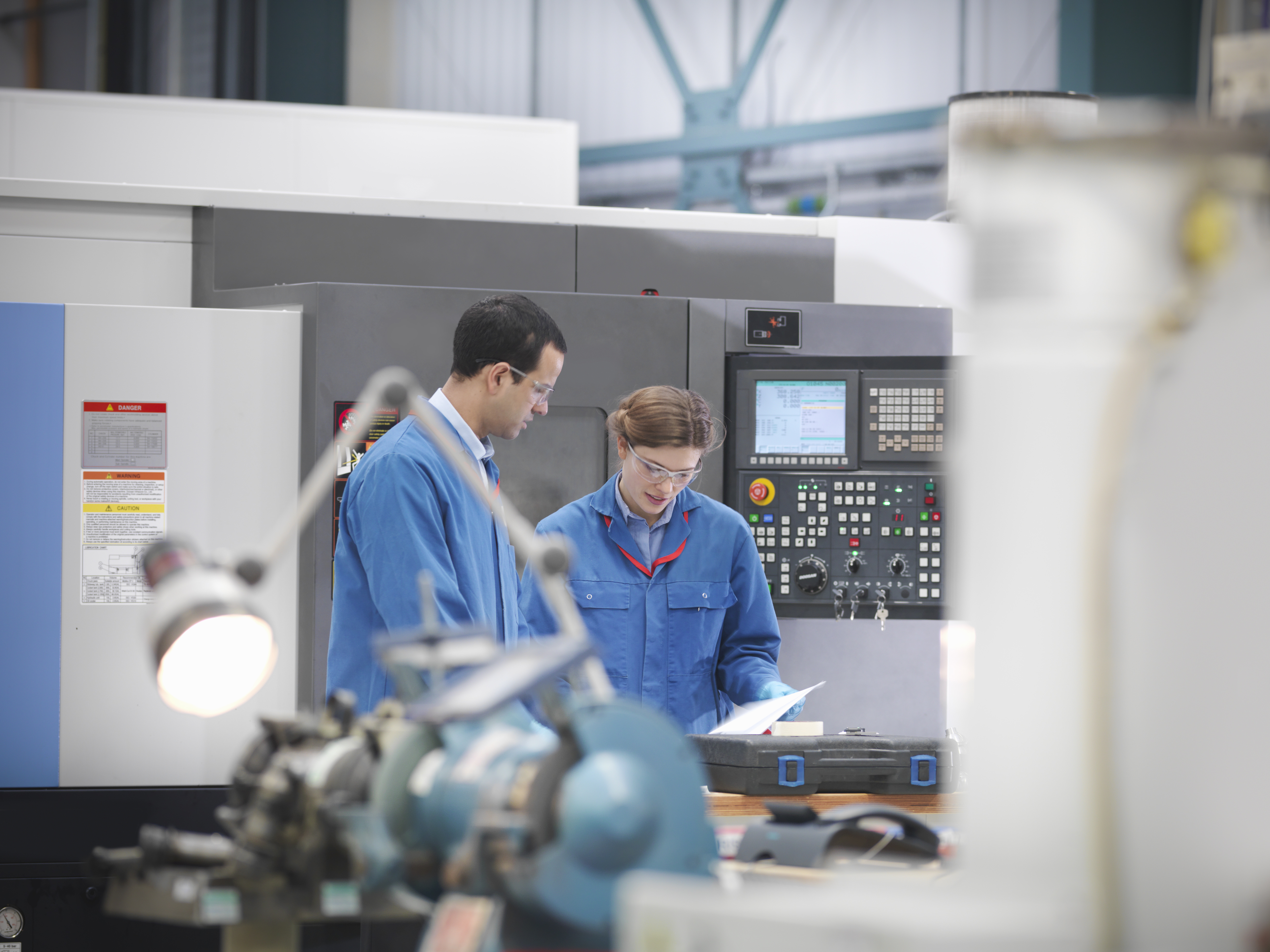 Two workers in blue uniforms operating CNC machinery in a modern manufacturing facility with industrial equipment.