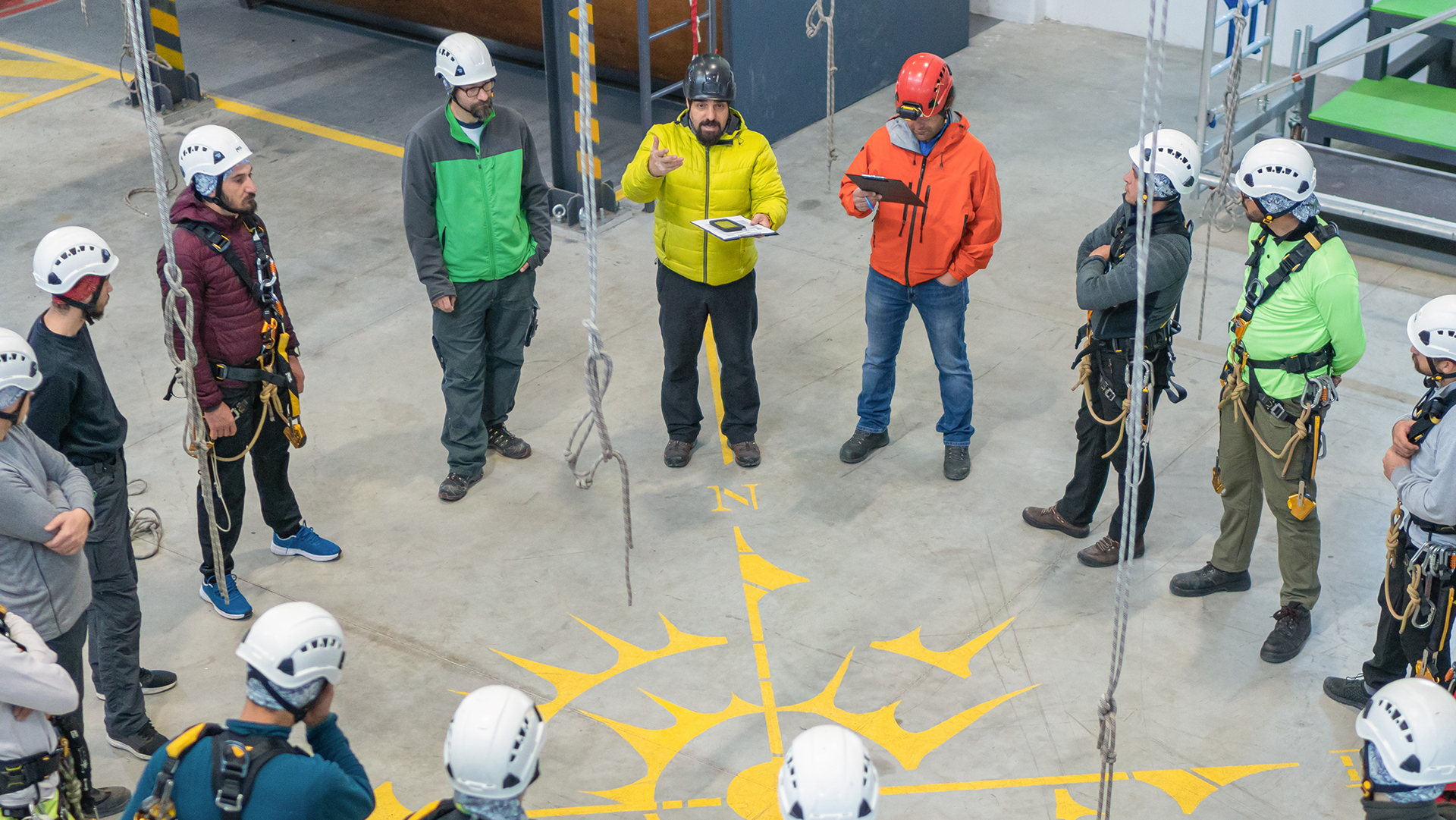 Safety training/fall protection session with workers in helmets and harnesses gathered in circle around instructor in yellow jacket.
