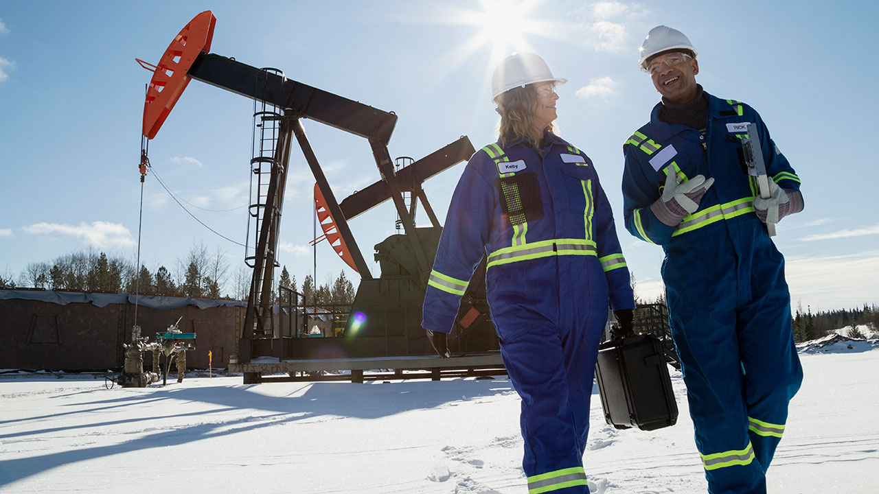 Two oilfield workers in blue safety suits and white hard hats walk across a snowy site near large pumpjack machinery under bright sunlight.