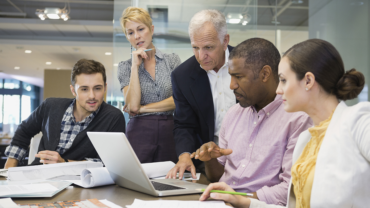 Diverse business team collaborating around a laptop in a modern office, reviewing documents and discussing project details.