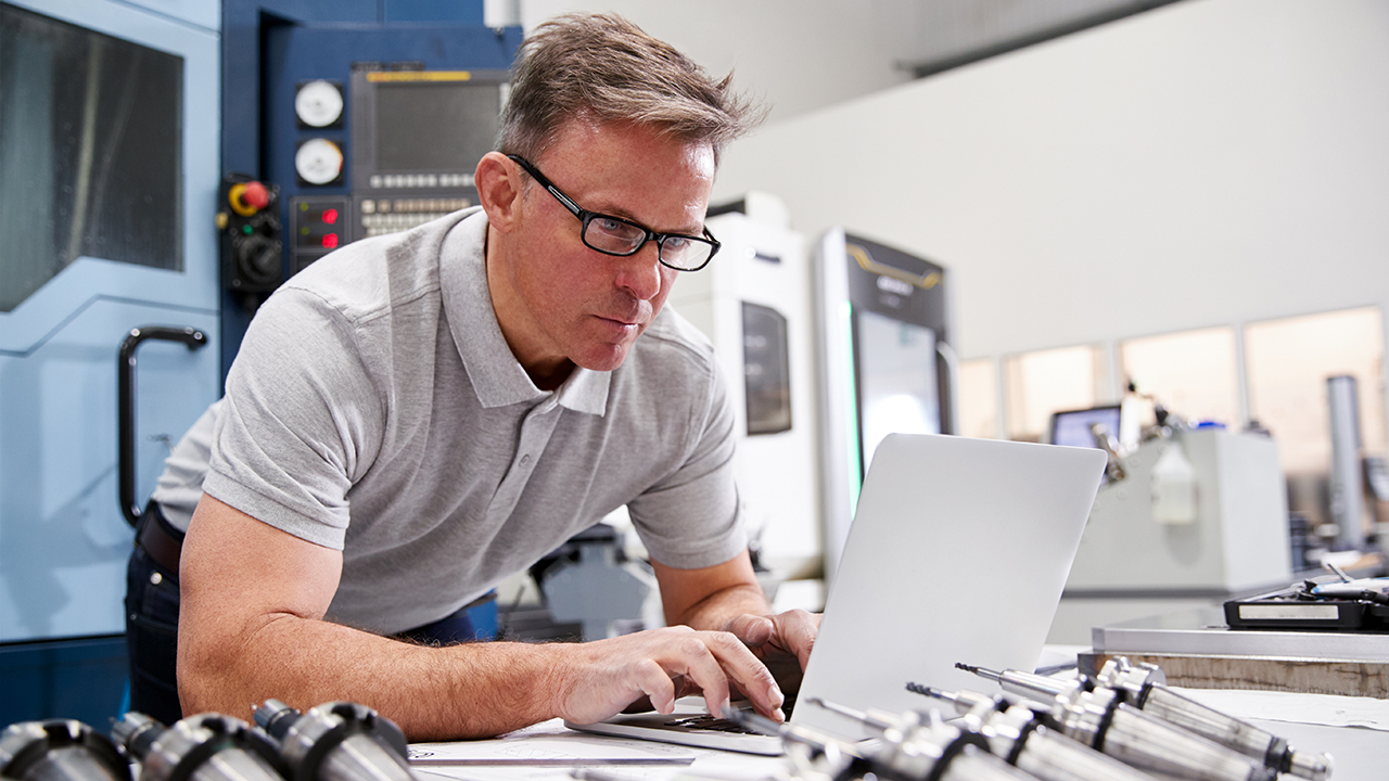 Man wearing glasses and gray polo shirt working on laptop in manufacturing facility with industrial machinery in background.