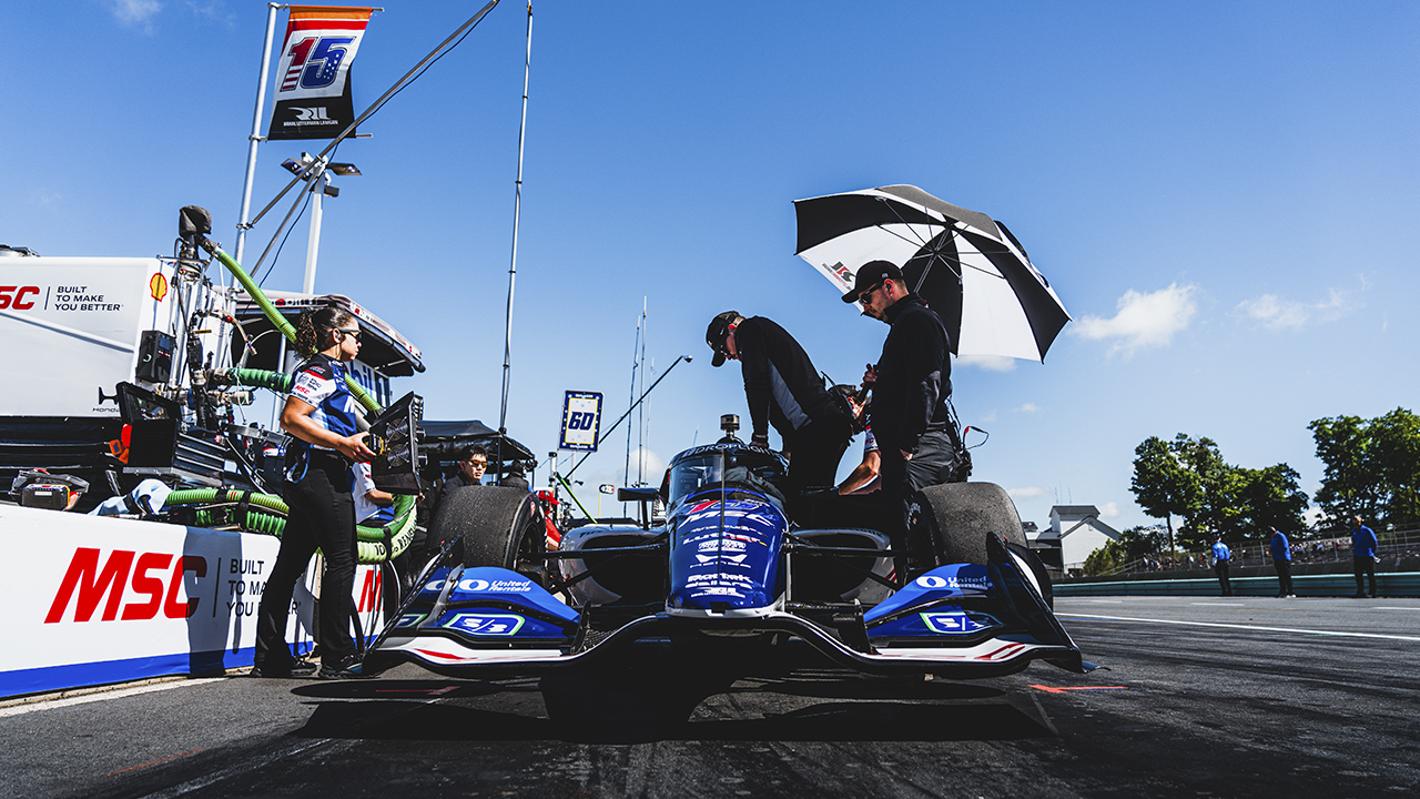 Racing crew working on blue MSC-branded IndyCar in pit lane under umbrella with clear blue sky and team equipment.