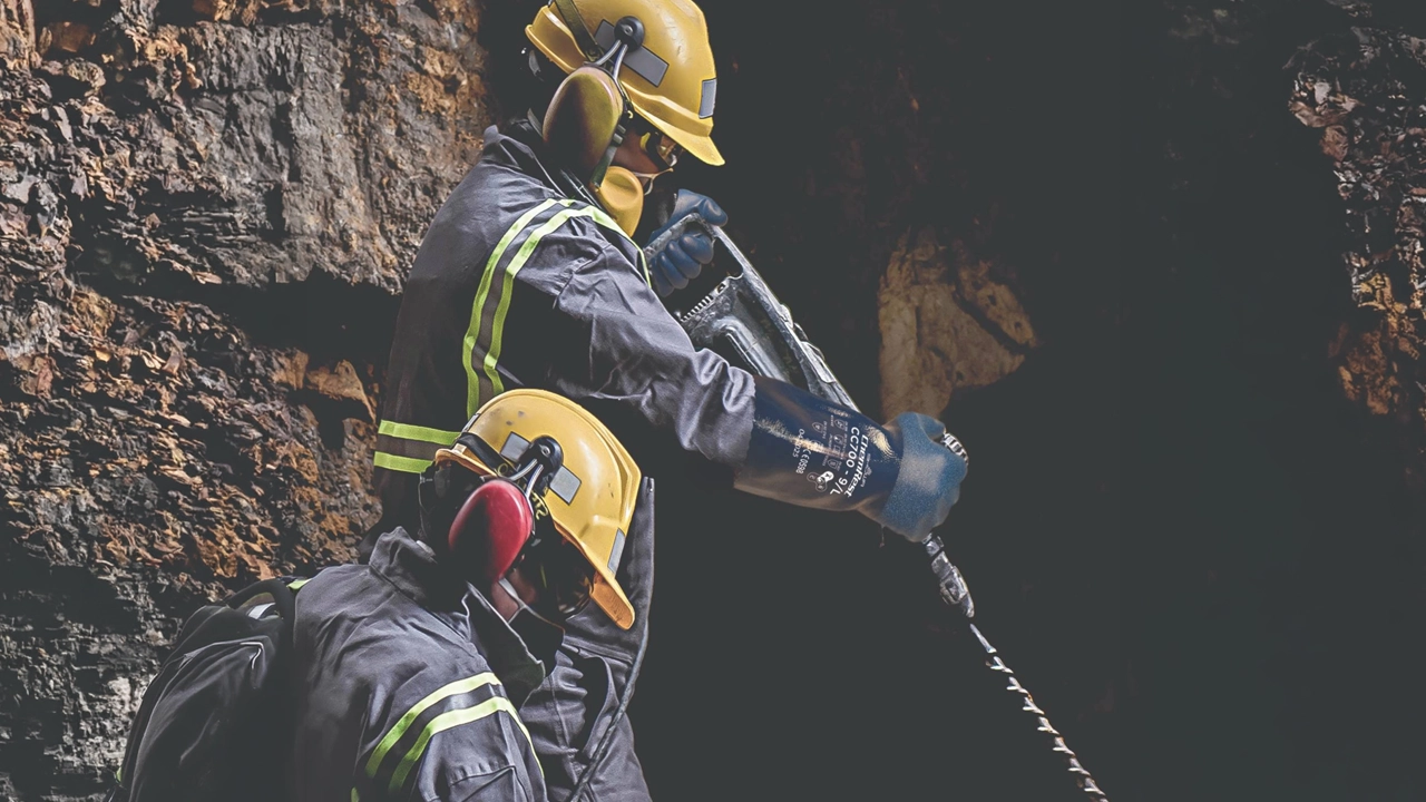 Two workers in yellow hard hats and black protective gear with reflective stripes using a drill in a smoky environment.
