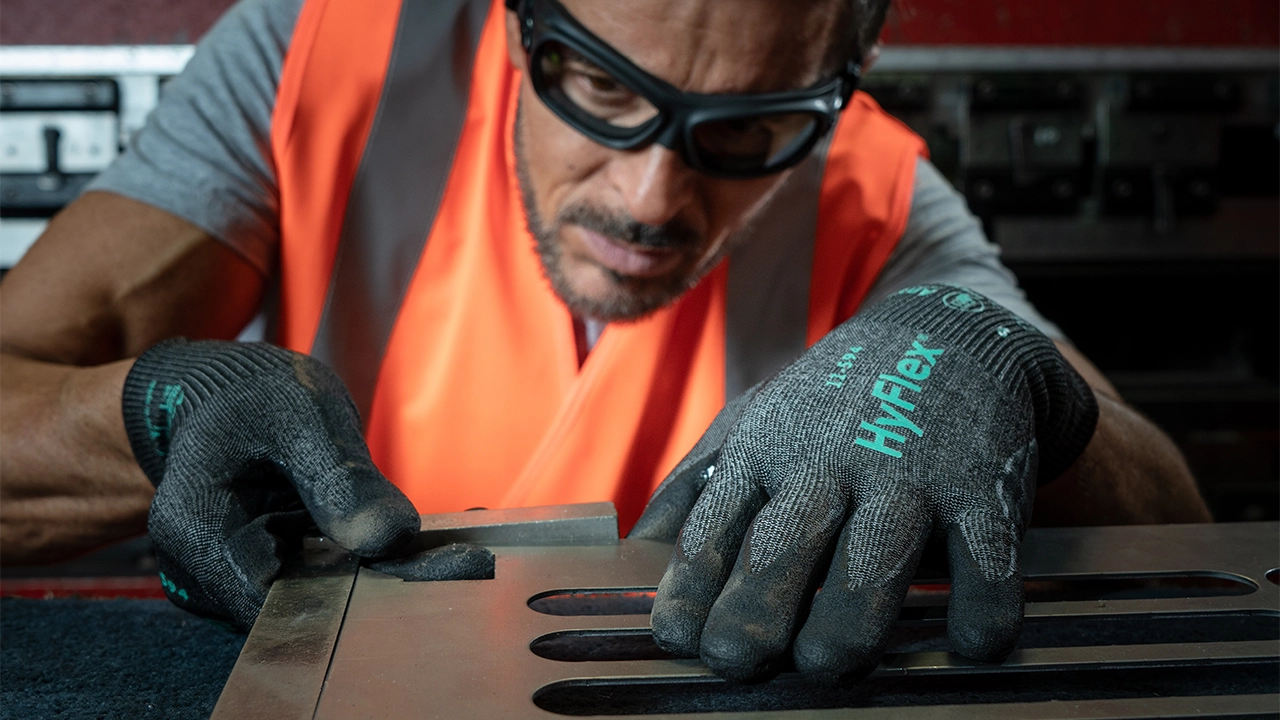 Worker wearing orange safety vest and black Hyflex work gloves using tool on metal surface in industrial setting