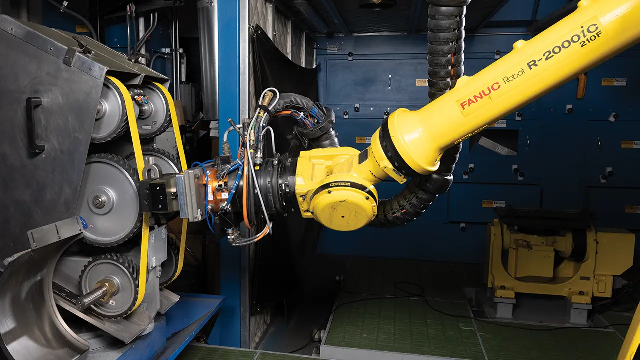Yellow FANUC R-2000iC robotic arm operating in industrial manufacturing cell with blue walls and metal machinery