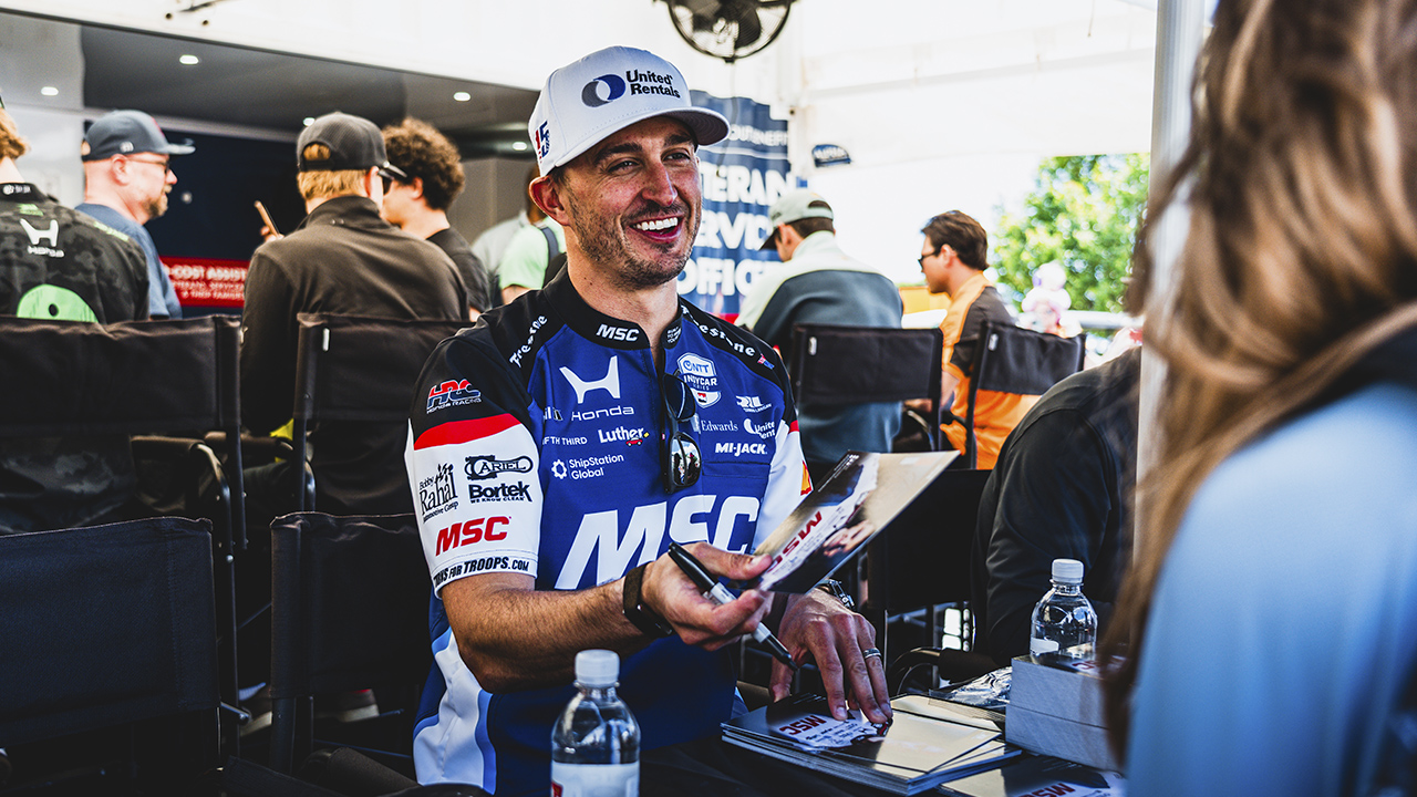 Race car driver, Graham Rahal, in blue and white MSC branded racing suit and cap smiling while signing autographs at event