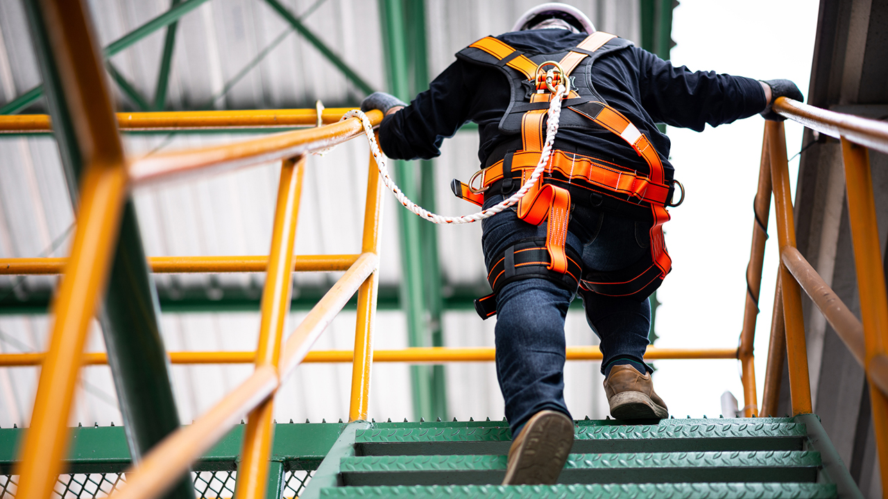 Worker in orange and black safety harness climbing industrial structure with yellow and green metal framework.