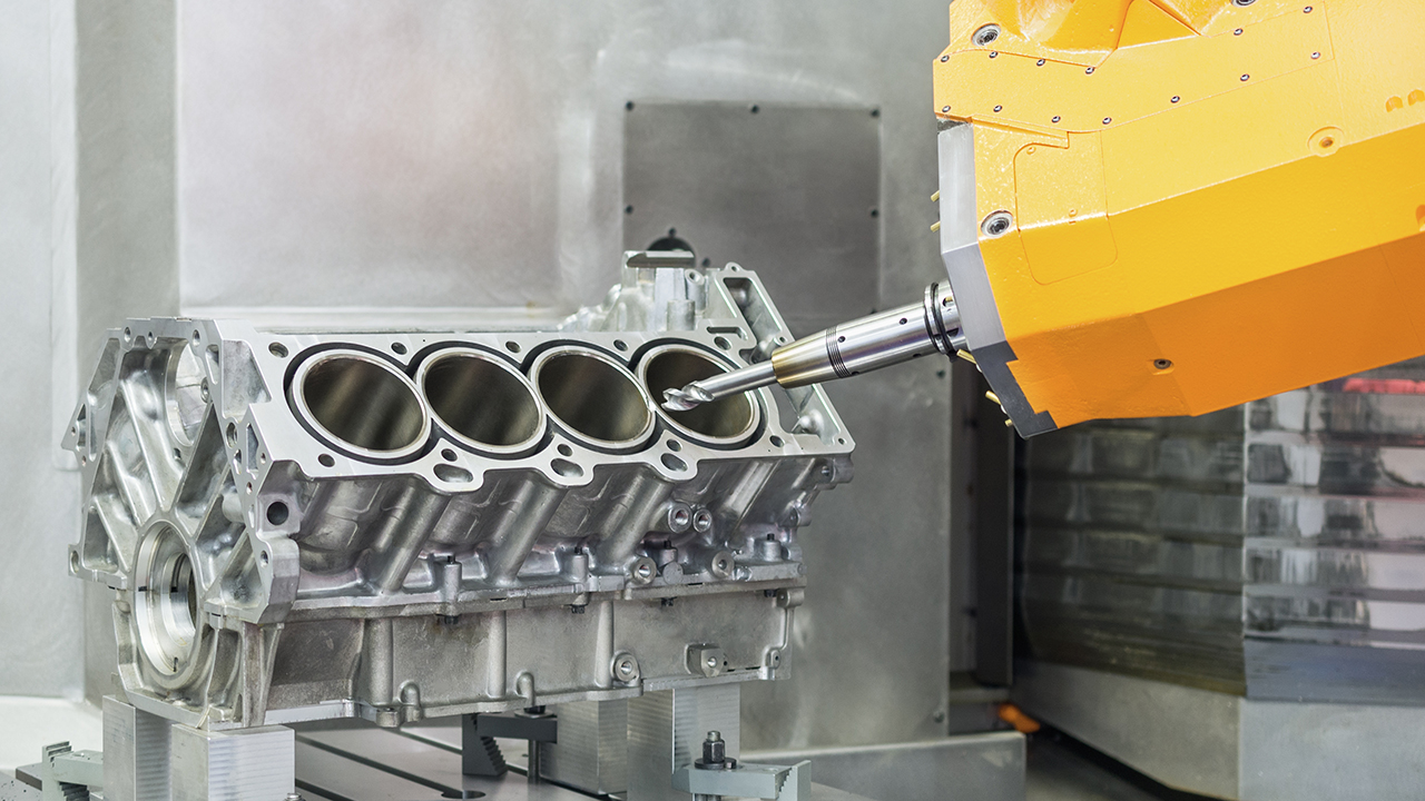 Yellow robotic arm with precision tool measuring or machining a silver automotive engine block in an industrial setting.