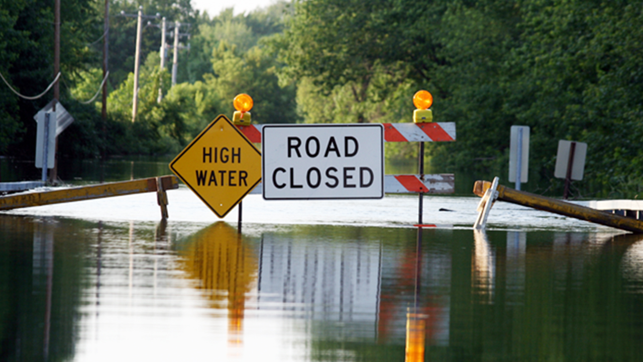 Flooded road with High Water and Road Closed signs, orange warning lights, and barricades surrounded by trees