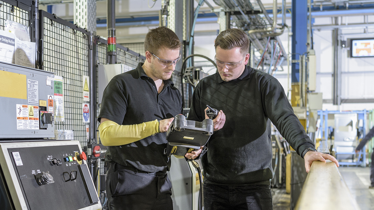 Two workers in safety glasses examining a handheld scanning device in an industrial manufacturing facility.
