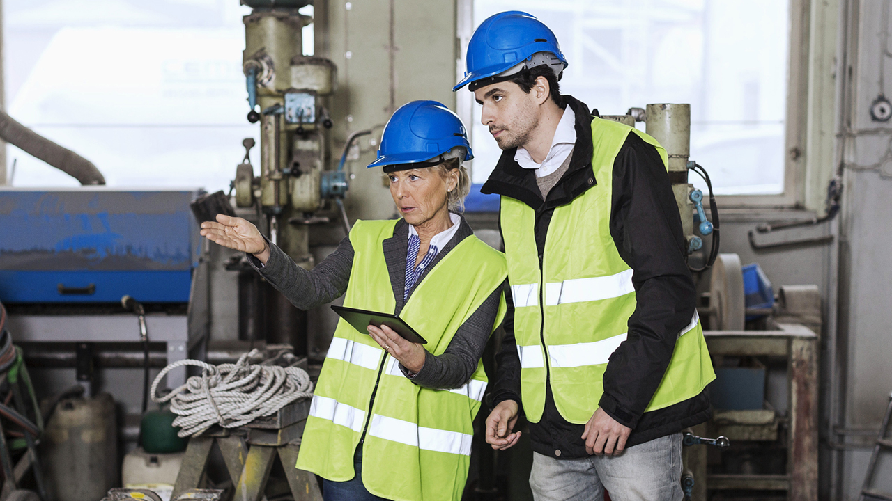 Two workers in blue hard hats and yellow safety vests discuss operations in an industrial facility with machinery.