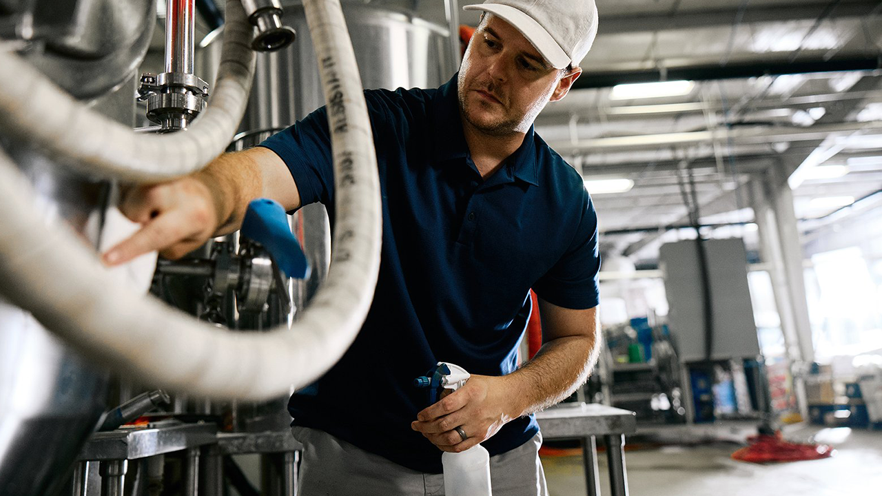 Worker cleaning industrial equipment with a cloth while holding a spray bottle in a manufacturing facility.