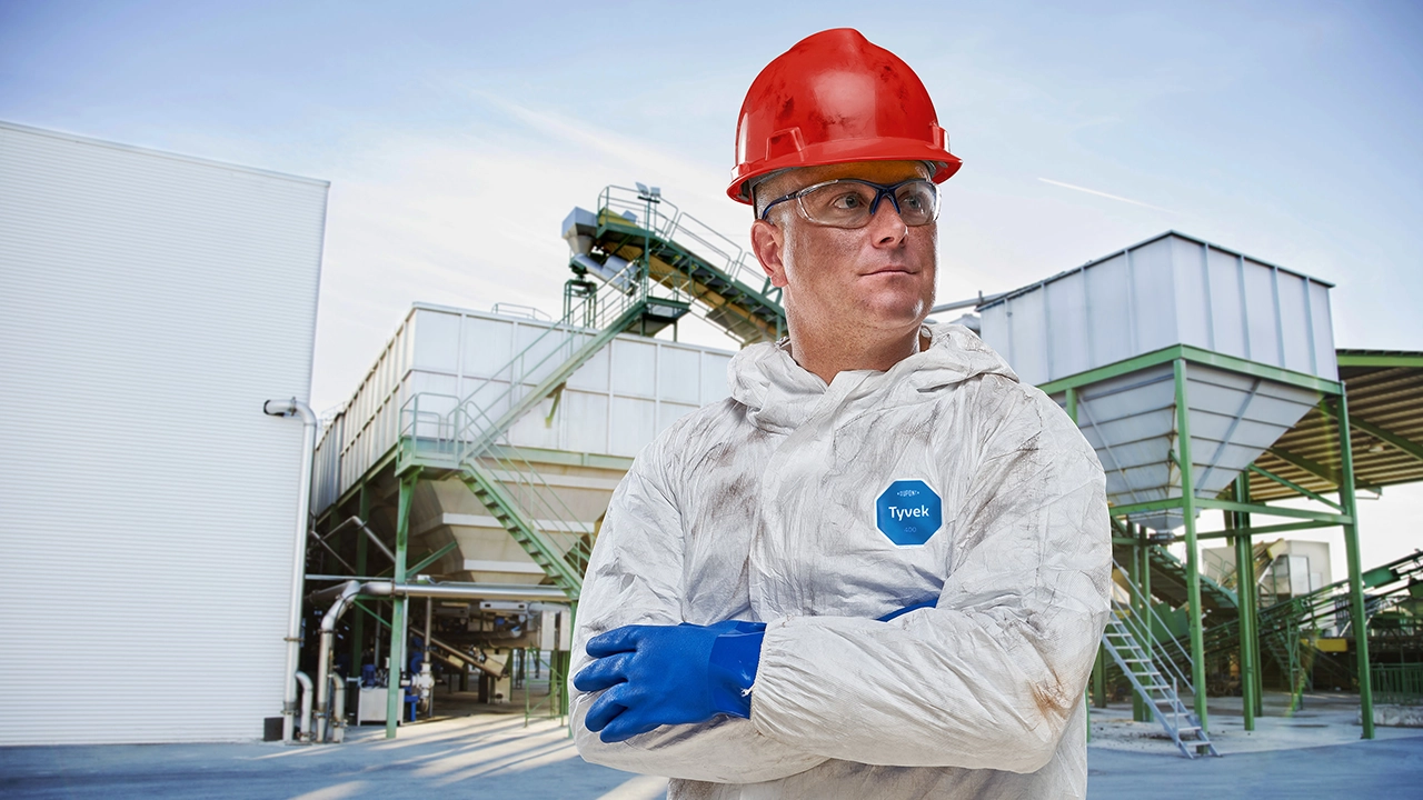 Worker in red hard hat, safety glasses, and white Tyvek protective suit with blue gloves at industrial facility