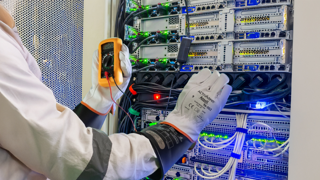 Technician in white coat using yellow Ansell multimeter to test network server rack with green and blue cables.