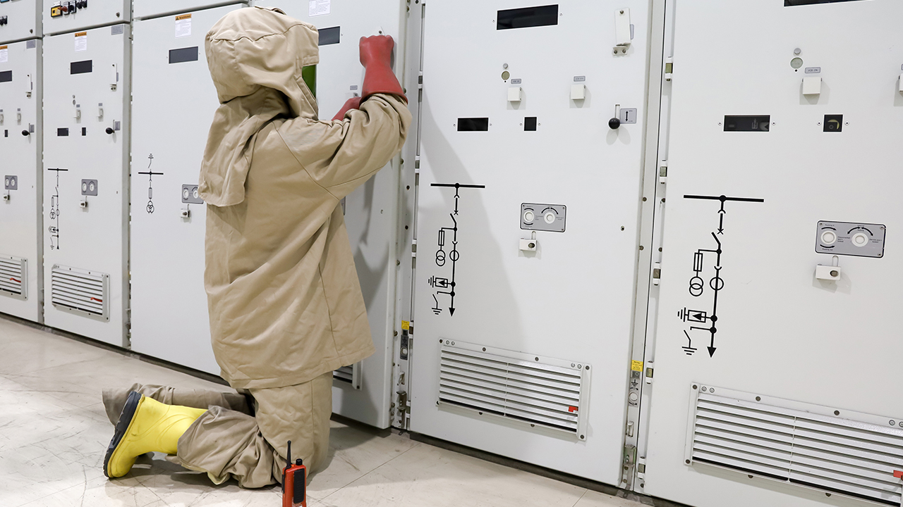 Worker in tan arc flash protective suit and hood operating electrical panel in industrial setting