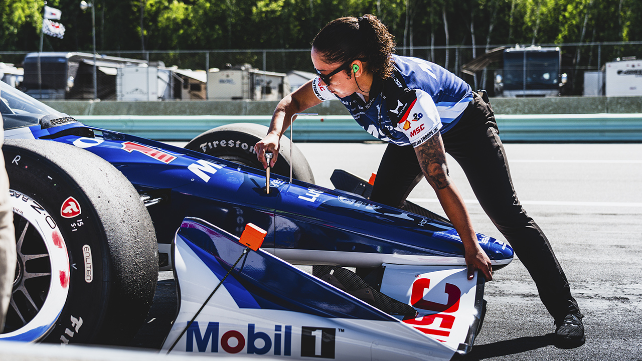 Female pit crew member in blue racing suit working on blue and white race car with Firestone tires and MSC logo on track