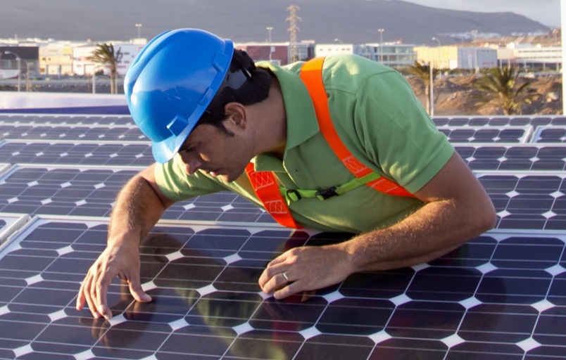 Two male workers in safety gear working on solar panels in a field with large wind turbines