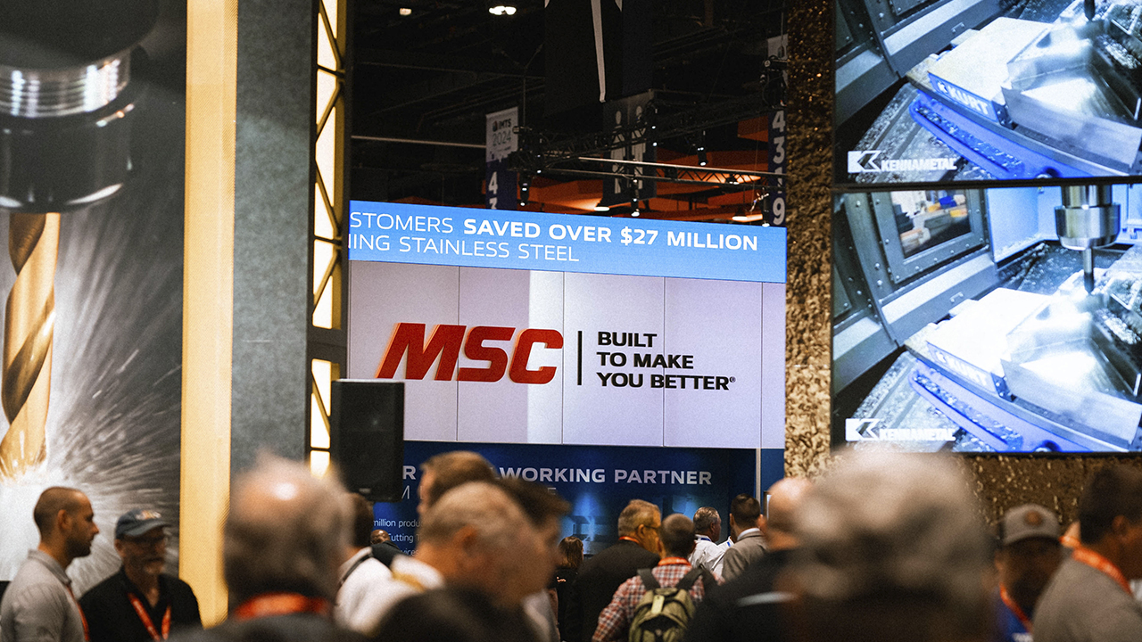Trade show booth with large MSC display screen showing customer savings message, surrounded by attendees in industrial setting.