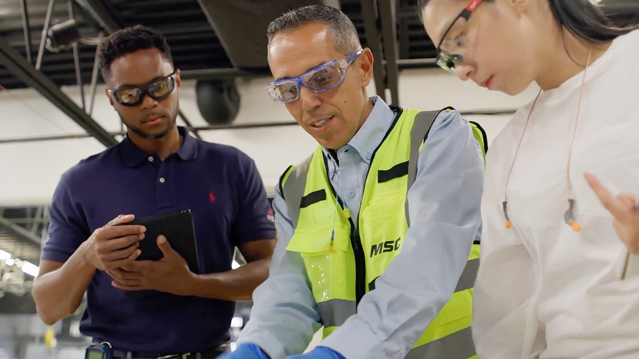 Three industrial workers in safety glasses collaborating in warehouse, one wearing MSC safety vest and blue gloves.