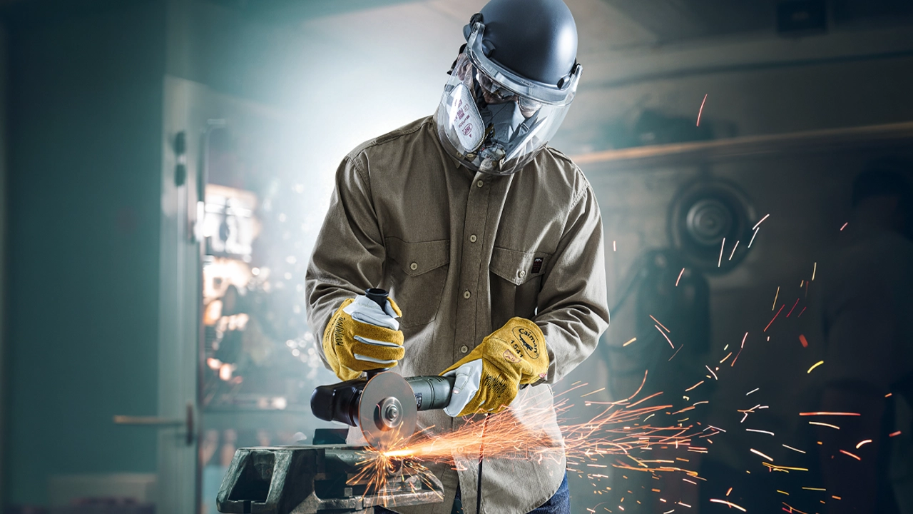 Worker in PIP protective helmet and respirator using angle grinder on metal, creating bright orange sparks in industrial setting.