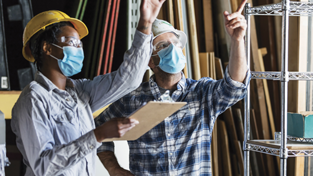 Two construction workers wearing hard hats review a clipboard as one gestures upward and the other points up inside a workshop with lumber and metal storage racks in the background.