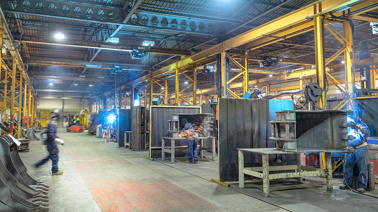 Industrial manufacturing facility with workers welding metal parts at workstations under yellow overhead crane system
