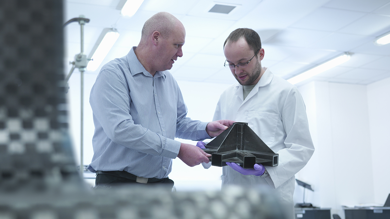 Two men in a manufacturing facility examining a black plastic component, one in business attire and one in a white lab coat.