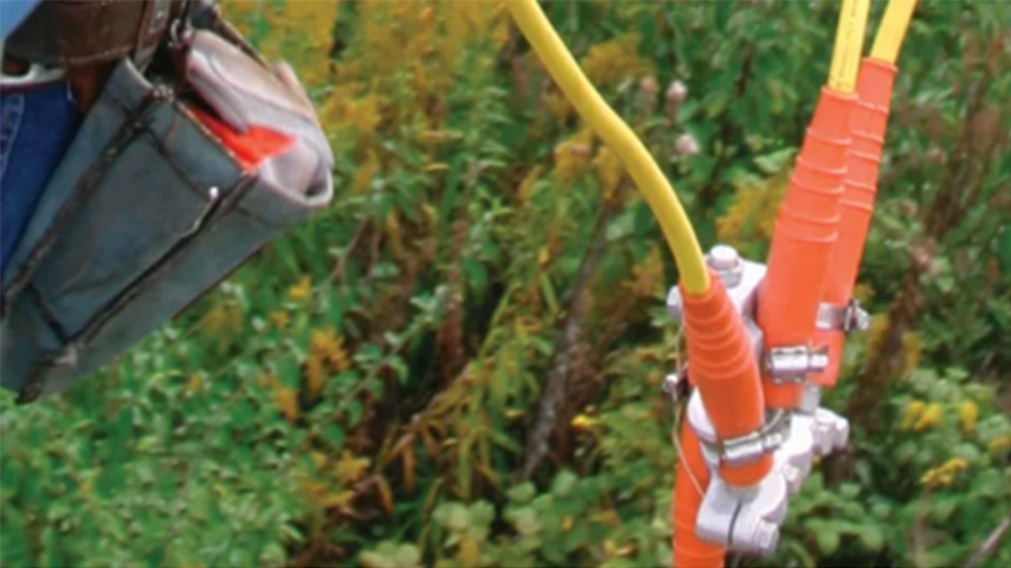 Close-up of insulated electrical grounding connectors attached to a metal clamp outdoors, with yellow cables extending over green vegetation in the background.
