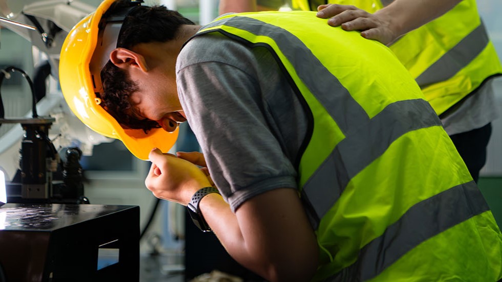 Worker in yellow hard hat and high-visibility vest being assisted by colleague after workplace injury or illness.
