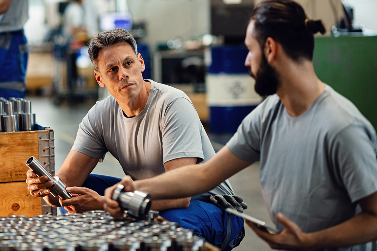 Two factory workers in gray shirts examining metal parts in an industrial workshop setting with machinery in background.