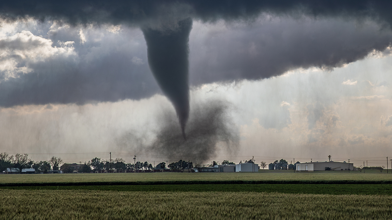 Tornado funnel cloud touching down in rural farmland with dark storm clouds, green fields, and buildings in distance.