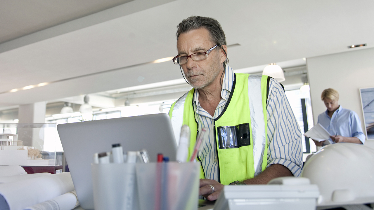 Man in safety vest and glasses working on laptop at desk in modern office with colleague in background