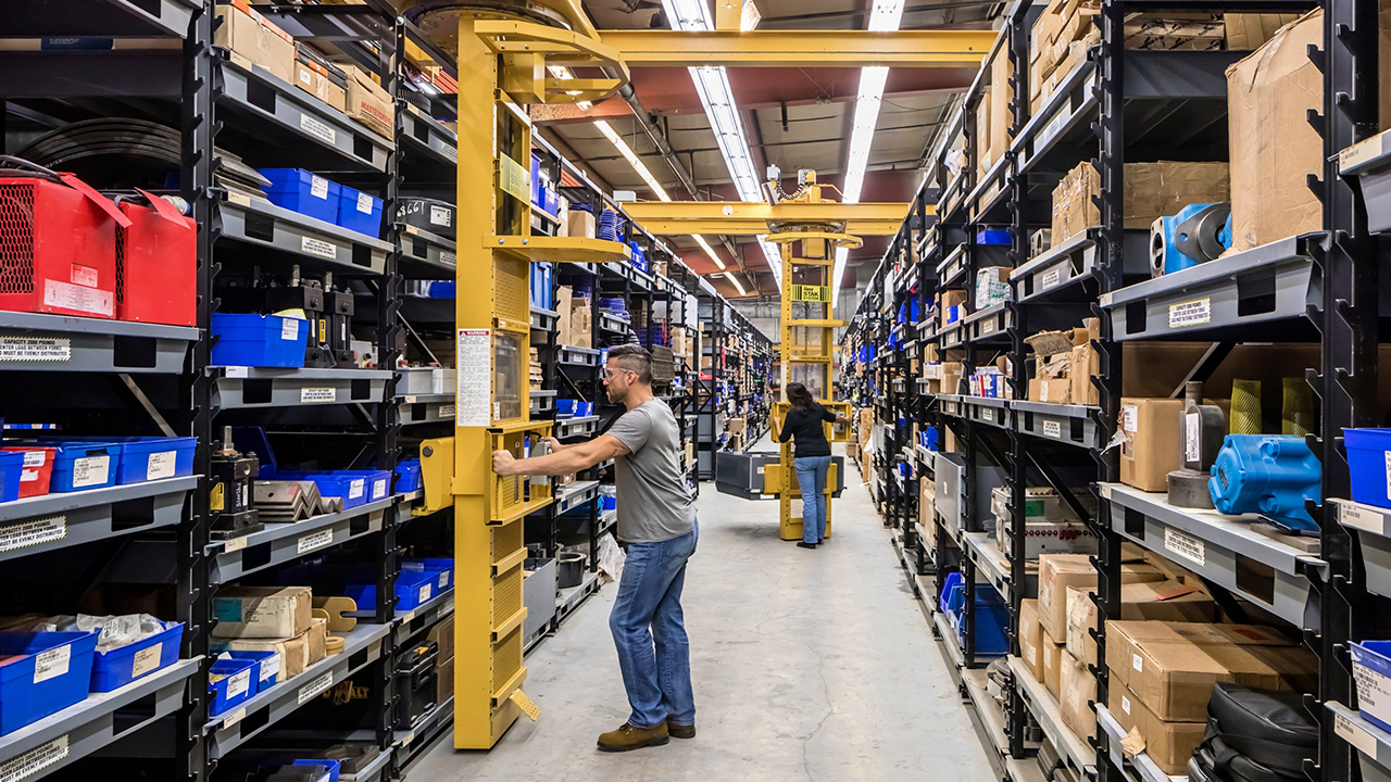 Warehouse worker operating yellow overhead crane in industrial storage aisle with black shelving and blue bins