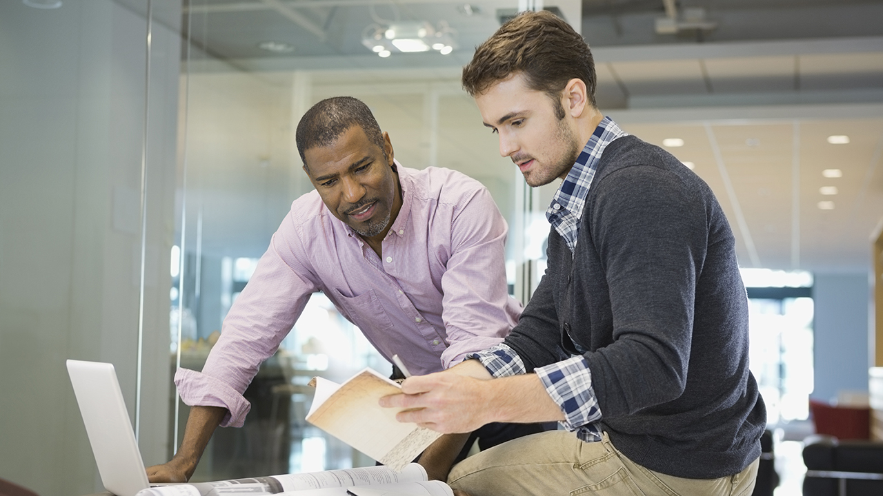 Two professionals collaborating in a modern office, reviewing notes and working on a laptop near a glass wall.