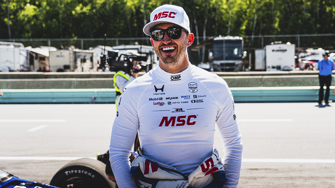 Graham Rahal, race car driver, wearing white MSC branded racing suit, cap, and sunglasses smiling at racetrack with RVs in background.