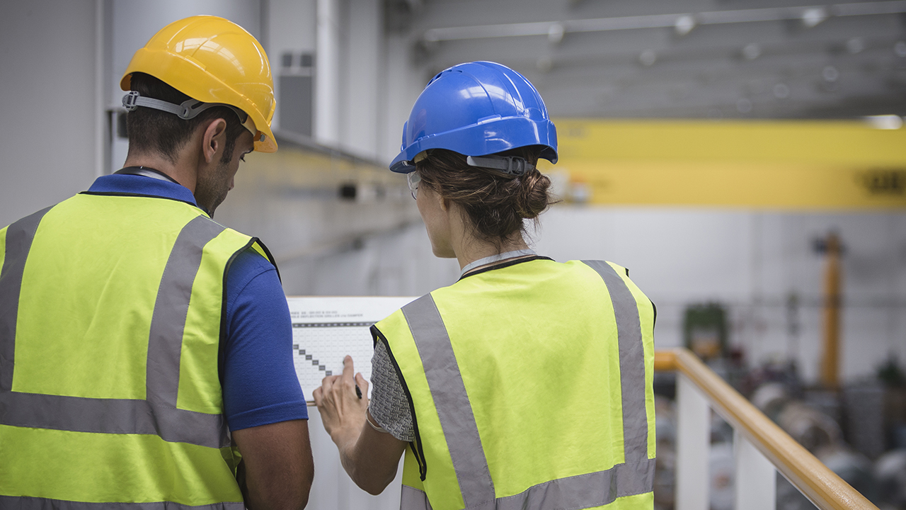 Two workers in yellow safety vests and hard hats, one yellow and one blue, reviewing documents in an industrial facility.