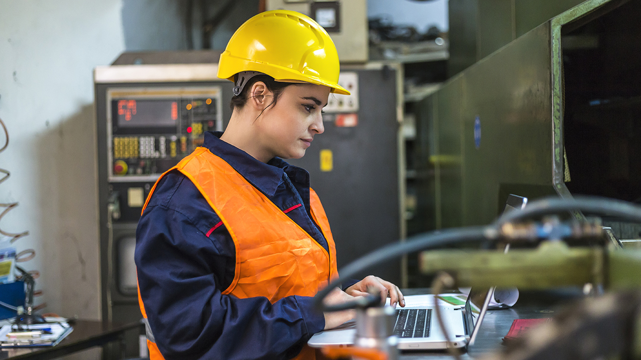 Female factory worker in yellow hard hat and orange safety vest using laptop near industrial machinery and control panel.