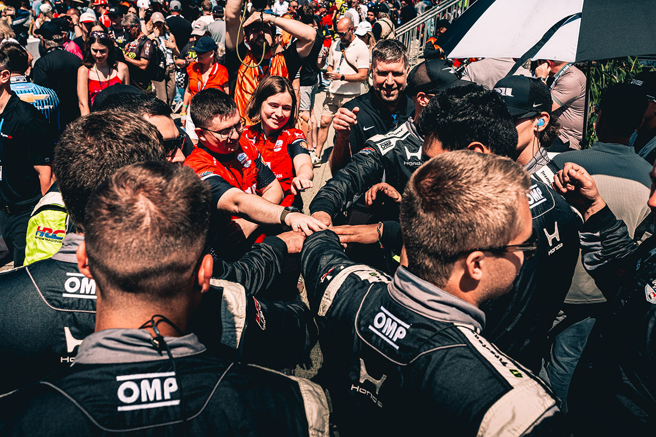 Racing team members in black OMP suits huddle together with fans in red and orange at crowded outdoor motorsport event