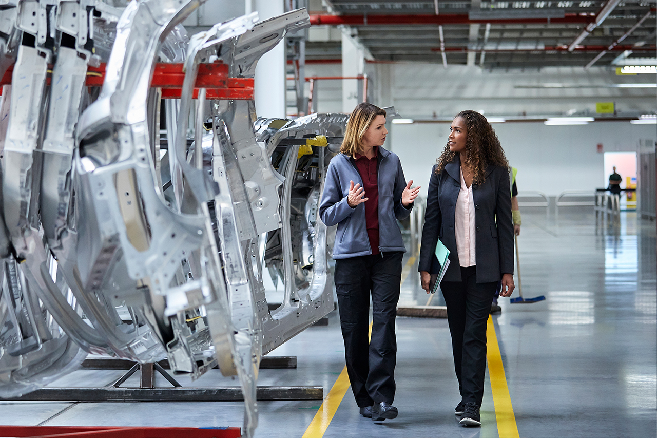Two women in business attire discussing work while walking past metal car frames in a modern automotive manufacturing facility.
